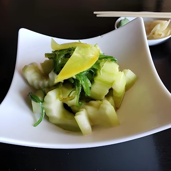 A white square bowl filled with light green pickled cucumbers and dark green seaweed salad.
